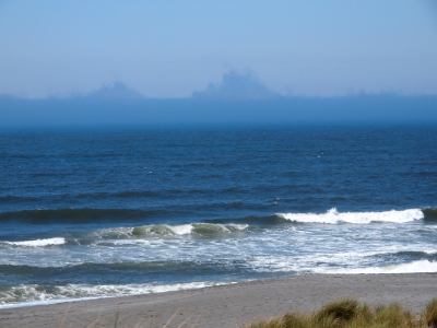 [View of the water with formations in the distant fog above the water line which appear to be small mountainous islands. There is white surf in the foreground leading to the sand.]